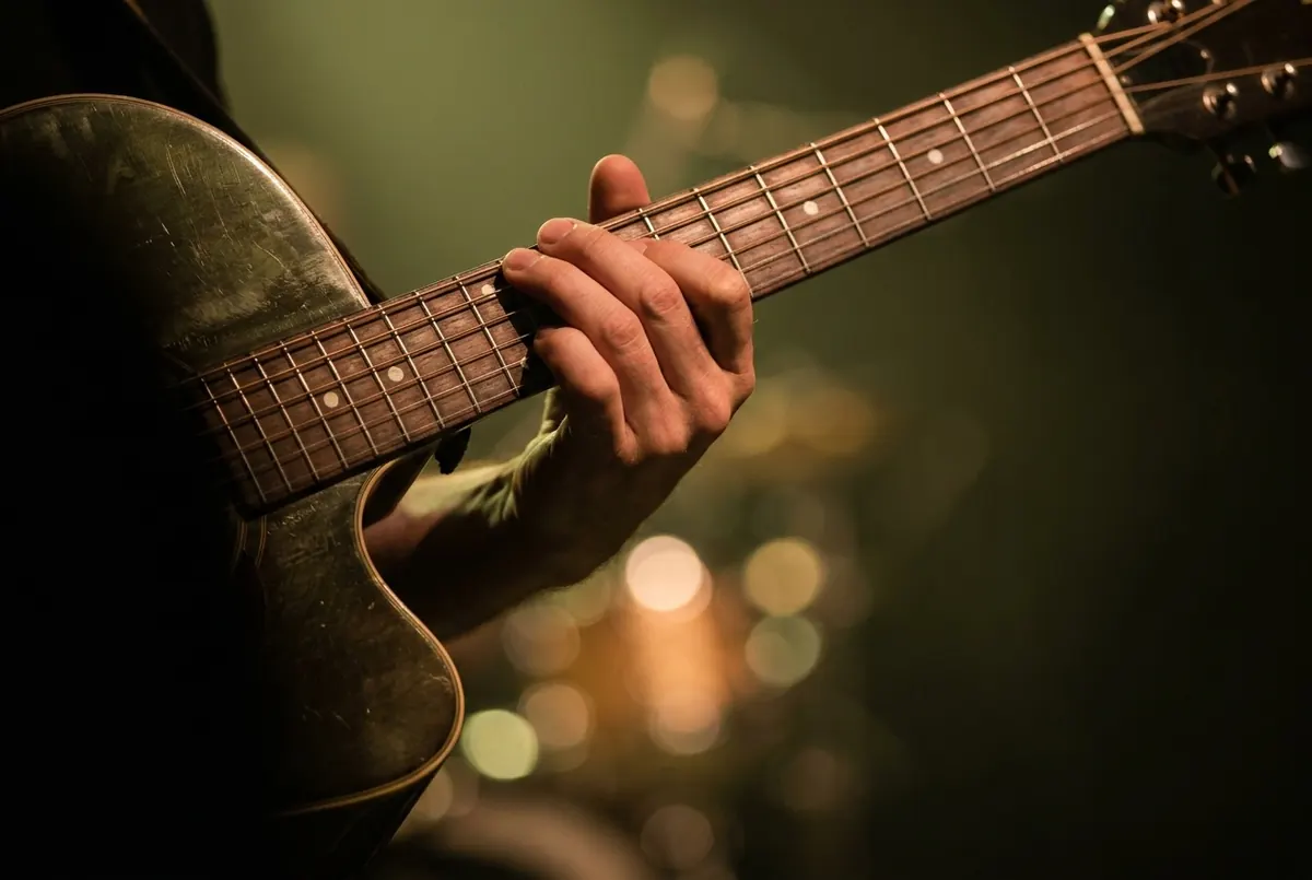 A hand fretting the neck of an acoustic guitar, lit in warm stage light